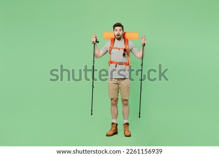 Full body shocked young traveler white man carry backpack stuff mat walk with trakking poles isolated on plain green background. Tourist leads active lifestyle. Hiking trek rest travel trip concept
