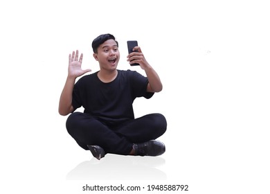 Full Body Shot Of Happy Asian Man In Black Clothes Sitting Isolated Against White Background. Attractive Teenager Holding Mobile Phone In Video Calling With His Hand Gesture Saying Hi