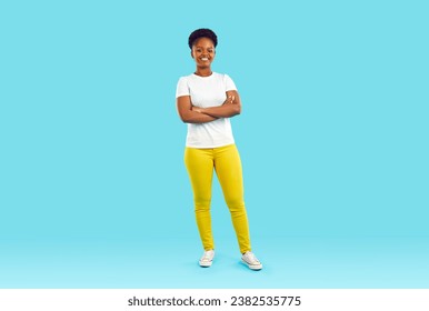 Full body shot of happy beautiful young African American woman in white Tshirt, yellow pants and comfortable shoes standing with arms folded isolated on blue color background. Casual fashion concept - Powered by Shutterstock