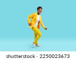 Full body shot of happy cheerful joyful beautiful young African American woman in bright yellow suit and white T shirt dancing isolated on blue color background. Fashion, party, having fun concept