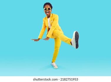 Full body shot of happy funny cheerful positive attractive young African American woman wearing stylish yellow suit and cool sunglasses dancing isolated on blue background. Party and fashion concept
