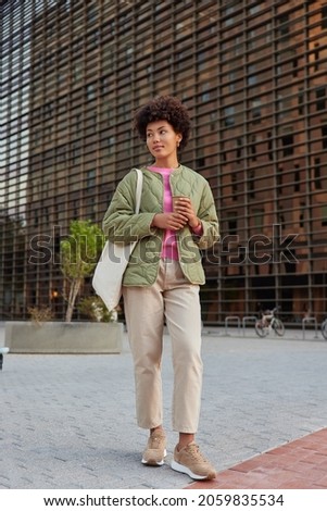 Full body shot of thoughtful young woman with curly hair dressed in stylish apparel holds paper disposable cup of coffee looks away carries fabric bag stands on pavement in city walks outside