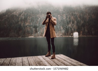 Full body shot of young male photographer in brown coat taking pictures with a small mirrorless camera standing on a lake house deck in lake plansee, Austria