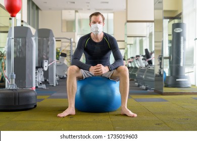 Full Body Shot Of Young Man With Mask Sitting On Exercise Ball At Gym During Corona Virus Covid-19