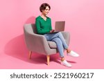 Full body shot of young programming woman freelancer sitting in cafeteria armchair using computer isolated on pink color background