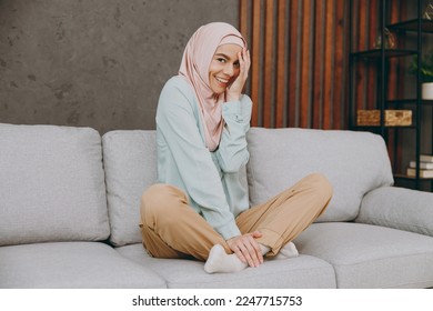 Full body shy calm fun young muslim woman wear hijab casual clothes sits on sofa couch stay at home flat rest relax spend free spare time in living room indoor. People uae middle eastern islam concept