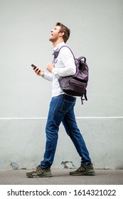 Full Body Side Portrait Smiling Man Walking With Mobile Phone And Bag By Gray Wall