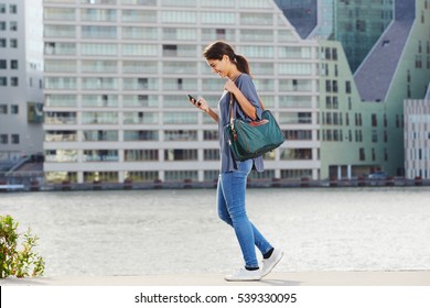 Full Body Side Portrait Of Young Woman Walking By The Water Using Cell Phone