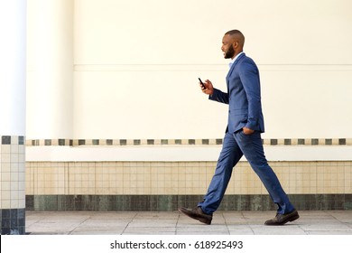 Full Body Side Portrait Of Young African Business Man Walking Outdoors With Mobile Phone