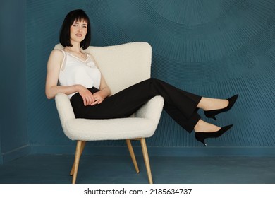 Full Body Side Profile View Of Smiling Young Brunette Woman Lifting Legs Arms Up Sitting And Relaxing On Chair On Blue Wall Background.Female In Black Pants, White Blouse And Classic High Heels Shoes.