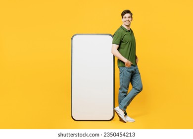 Full body side profile view young happy man he wears green t-shirt casual clothes stand near big huge blank screen mobile cell phone smartphone with workspace area isolated on plain yellow background - Powered by Shutterstock