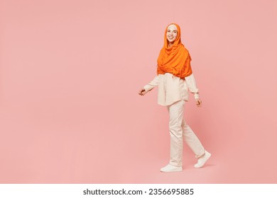 Full body side profile view smiling young arabian asian muslim woman wear orange abaya hijab walk go strolling look camera isolated on plain pink background. Uae middle eastern islam religious concept - Powered by Shutterstock