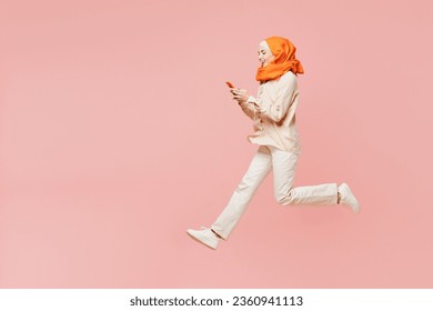 Full body side profile view young arabian asian muslim woman wearing orange abaya hijab jump high use mobile cell phone isolated on plain pink background. Uae middle eastern islam religious concept - Powered by Shutterstock