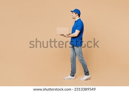 Full body side profile view delivery guy employee man wear blue cap t-shirt uniform workwear work as dealer courier hold cardboard box go walk isolated on plain light beige background. Service concept