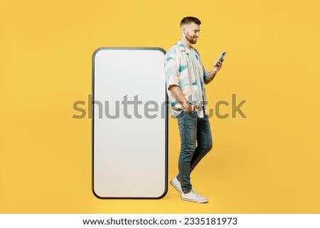 Full body side profile view young man he wearing blue shirt white t-shirt casual clothes big huge blank screen mobile cell phone with area using smartphone isolated on plain yellow background studio