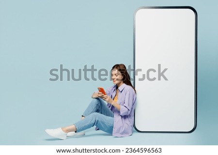 Full body side profile view young woman wears purple shirt yellow t-shirt casual clothes sit big huge blank screen mobile cell phone with area use smartphone isolated on plain light blue background