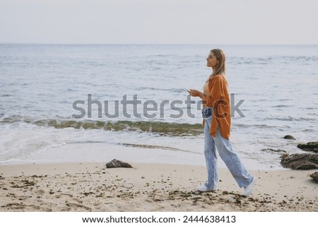 Full body side profile view young woman she wears orange shirt casual clothes hold use mobile cell phone strolling walk on sea ocean sand shore beach outdoor seaside in summer day. Lifestyle concept