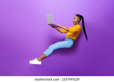 Full body side view of amazed young African American woman levitating while browsing netbook computer on purple background in light studio