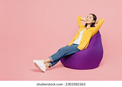 Full body side view calm young woman of Asian ethnicity wear yellow shirt white t-shirt sit in bag chair hold hands behind neck isolated on plain pastel light pink background studio. Lifestyle concept