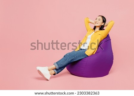 Full body side view calm young woman of Asian ethnicity wear yellow shirt white t-shirt sit in bag chair hold hands behind neck isolated on plain pastel light pink background studio. Lifestyle concept