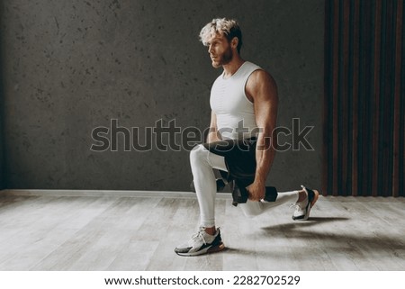 Full body side view caucasian young strong sporty athletic sportsman man wear white tank shirt black shorts lifting hold dumbbells do squat lunges warm up training indoor at gym. Workout sport concept