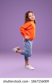 Full Body Side View Of Cheerful Little Girl In Bright Orange Sweatshirt And Denim Pants Standing On One Leg Against Purple Background