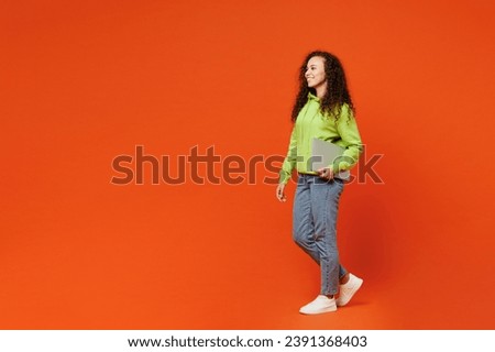 Full body side view confident young IT woman of African American ethnicity wears green hoody casual clothes hold closed laptop pc computer isolated on plain red orange background. Lifestyle concept