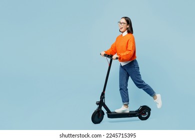 Full body side view cool happy young woman of Asian ethnicity wear orange sweater glasses riding electric scooter isolated on plain pastel light blue cyan background studio People lifestyle concept