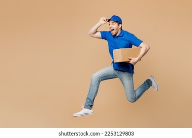 Full body side view delivery guy employee man wear blue cap t-shirt uniform workwear work as dealer courier jump high hold cardboard box run isolated on plain light beige background. Service concept - Powered by Shutterstock