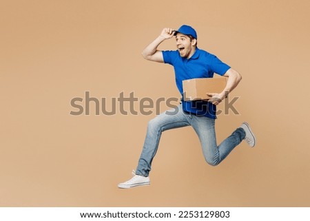 Full body side view delivery guy employee man wear blue cap t-shirt uniform workwear work as dealer courier jump high hold cardboard box run isolated on plain light beige background. Service concept