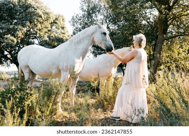 Full body side view of elderly gray haired horsewoman feeding gray horse from palm while standing in countryside near green trees on sunny day - Powered by Shutterstock