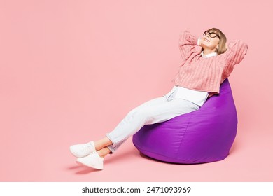 Full body side view elderly woman 50s years old wear sweater shirt casual clothes glasses sit in bag chair hold hands behind neck isolated on plain pink background studio portrait. Lifestyle concept