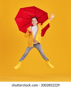 Full body side view of excited little girl in yellow raincoat and boots with red umbrella having fun and jumping above ground against yellow background