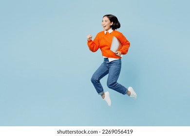 Full body side view excited young IT woman of Asian ethnicity wear orange sweater glasses jump high hold closed laptop pc computer run fast isolated on plain pastel light blue cyan background studio