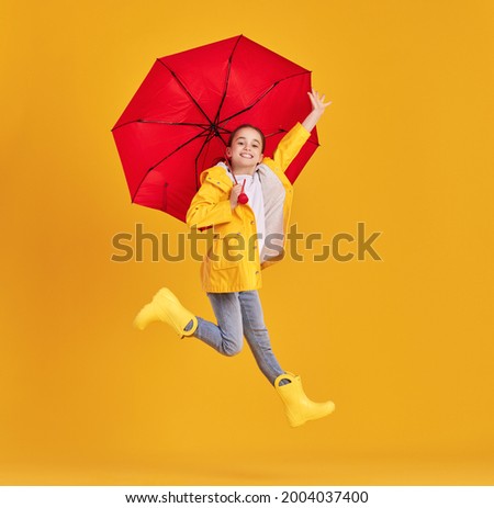 Full body side view of excited little girl in yellow raincoat and boots with red umbrella having fun and jumping above ground against yellow background