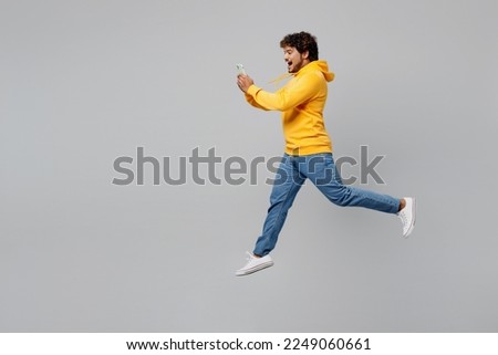 Full body side view excited young Indian man 20s he wearing casual yellow hoody jump high hold in hand mobile cell phone isolated on plain grey background studio portrait. People lifestyle portrait