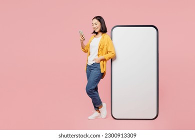 Full body side view fun young woman of Asian ethnicity wear yellow shirt white t-shirt big huge blank screen mobile cell phone with area use smartphone isolated on plain pastel light pink background - Shutterstock ID 2300913309