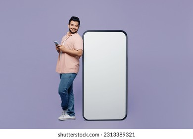 Full body side view fun young Indian man he wear pink shirt white t-shirt casual clothes big huge blank screen mobile cell phone with area using smartphone isolated on plain pastel purple background - Shutterstock ID 2313389321