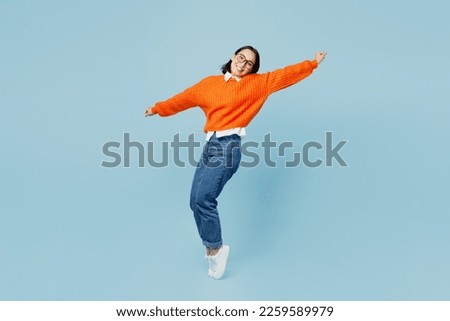 Full body side view fun young woman of Asian ethnicity wear orange sweater glasses stand on toes leaning back with outstretched hands dance isolated on plain pastel light blue cyan background studio