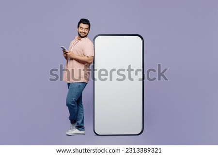 Full body side view fun young Indian man he wear pink shirt white t-shirt casual clothes big huge blank screen mobile cell phone with area using smartphone isolated on plain pastel purple background