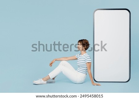 Full body side view fun young woman wears striped t-shirt casual clothes sitting near big huge blank screen mobile cell phone smartphone with area isolated on plain blue background. Lifestyle concept