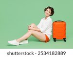 Full body side view happy traveler woman wear casual clothes hold sit near suitcase bag look camera isolated on plain green background. Tourist travel abroad in free time rest. Air flight trip concept