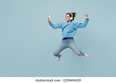 Full body side view happy young woman wear knitted sweater headphones listen to music jump high hold in hand use mobile cell phone isolated on plain pastel light blue cyan background studio portrait