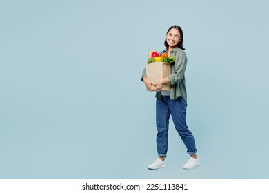 Full body side view happy young woman in casual clothes hold brown paper bag with food products look camera isolated on plain blue background studio portrait. Delivery service from shop or restaurant