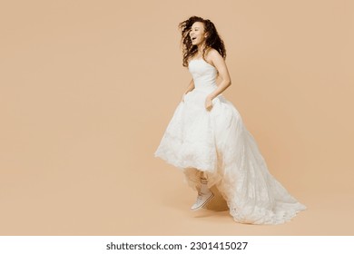 Full body side view happy young woman bride wears wedding dress posing lifts her dress show sneakers jump high isolated on plain beige background studio portrait. Ceremony celebration party concept