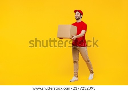Full body side view happy delivery guy employee man in red cap T-shirt uniform workwear work as dealer courier hold cardboard box isolated on plain yellow background studio portrait. Service concept