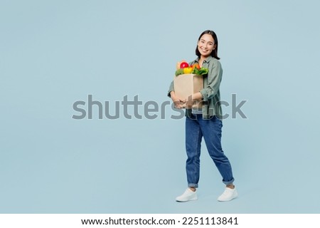 Full body side view happy young woman in casual clothes hold brown paper bag with food products look camera isolated on plain blue background studio portrait. Delivery service from shop or restaurant