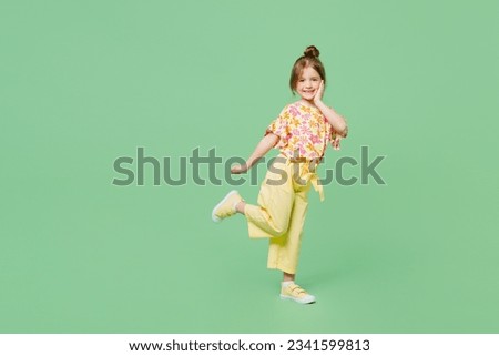 Full body side view little child kid girl 6-7 years old wears casual clothes look camera raise up leg hold face isolated on plain green background studio. Mother's Day love family lifestyle concept
