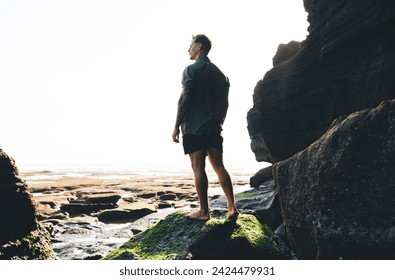 Full body side view of male traveler standing on rock covered with moss and overlooking vast expanse of tranquil sea - Powered by Shutterstock