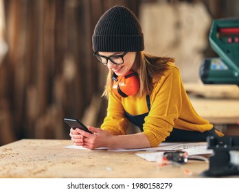 Full body side view of positive young female woodworker in apron messaging on mobile phone while standing near workbench in joinery workshop - Powered by Shutterstock